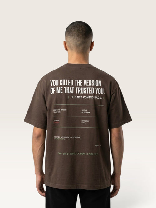 Coffee - A man viewed from behind wearing a dark brown t-shirt with a large white graphic text print on the back, paired with black pants, against a light beige studio background.