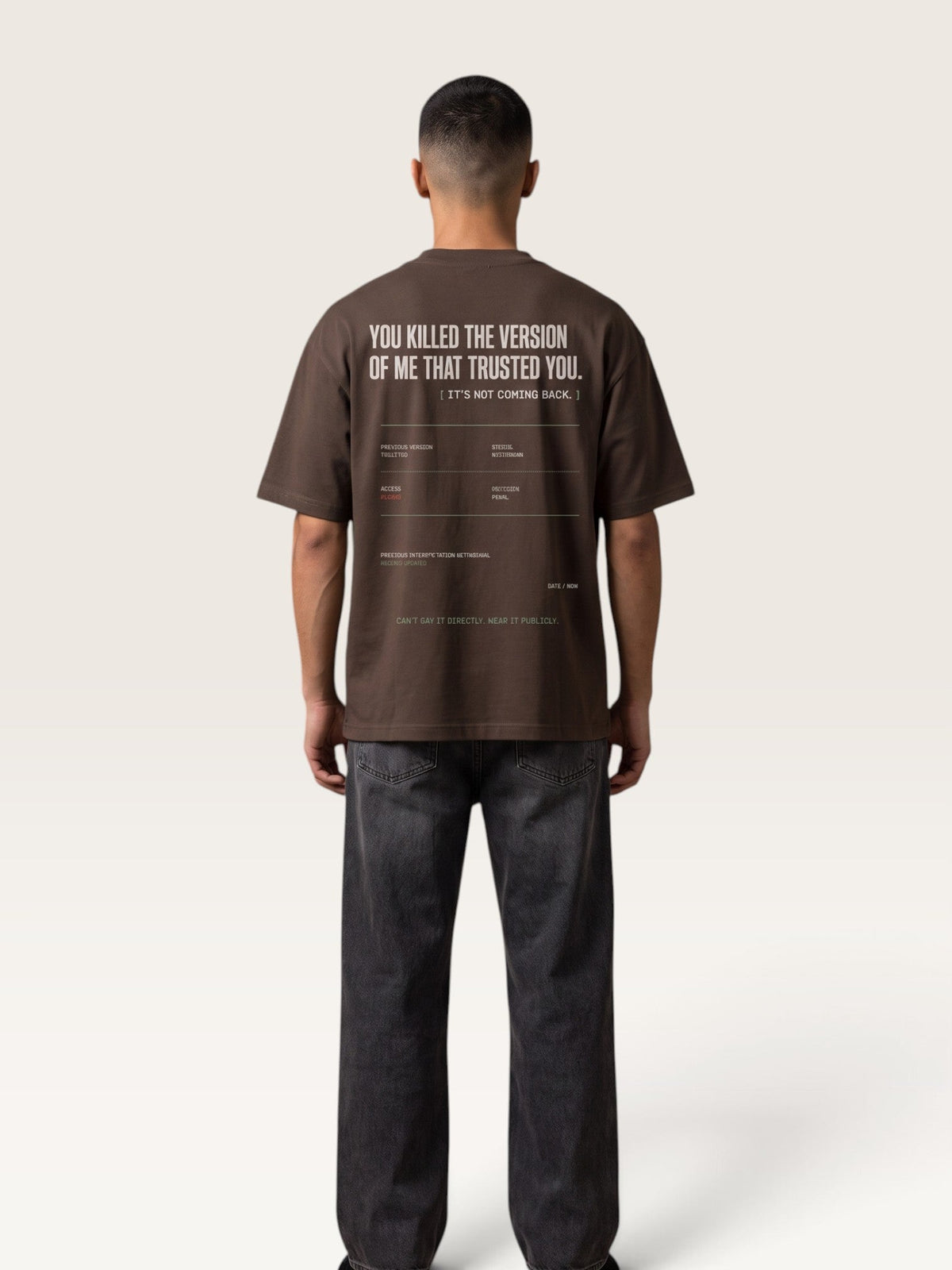 Coffee - A man viewed from behind wearing a dark brown t-shirt with white graphic text on the back and dark gray jeans, standing against a light beige studio background.