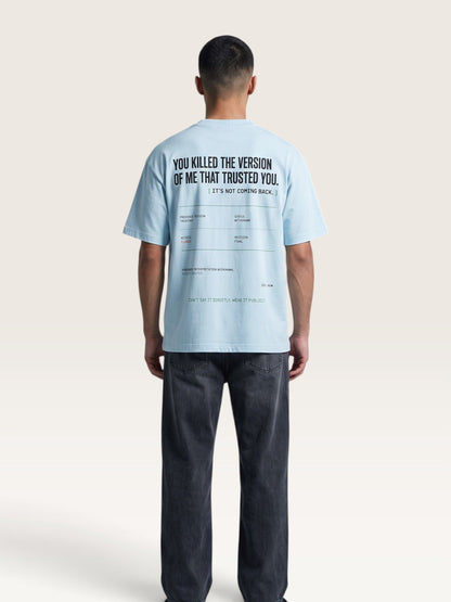 Sky Blue - A man viewed from behind wearing a light blue graphic t-shirt and dark gray jeans against a plain off-white studio background.