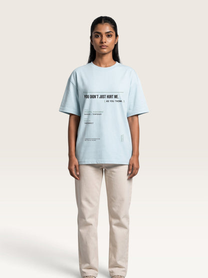 Sky Blue - A model stands against a light beige studio background wearing an oversized light blue graphic T-shirt and cream-colored trousers. The T-shirt features black printed text on the chest reading 'YOU DON'T JUST HURT ME.