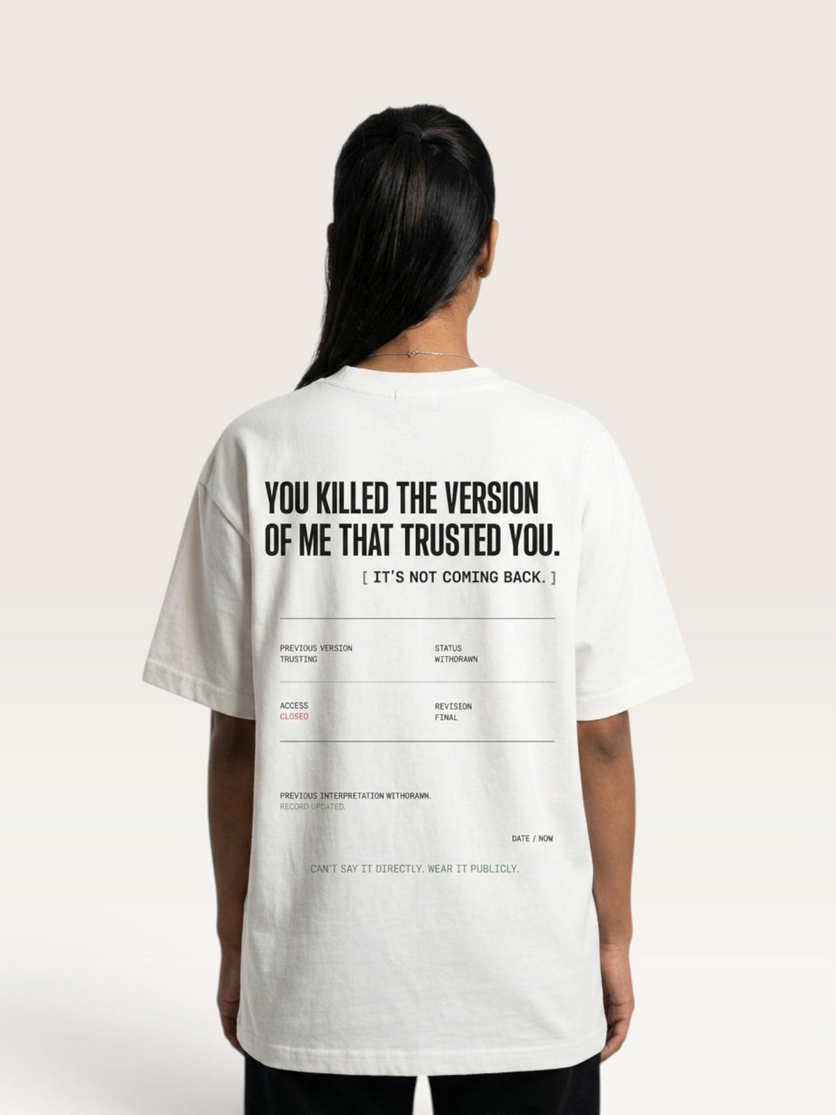 White - woman viewed from behind wearing an oversized white graphic T-shirt with black text on the back, set against a plain light beige studio background.